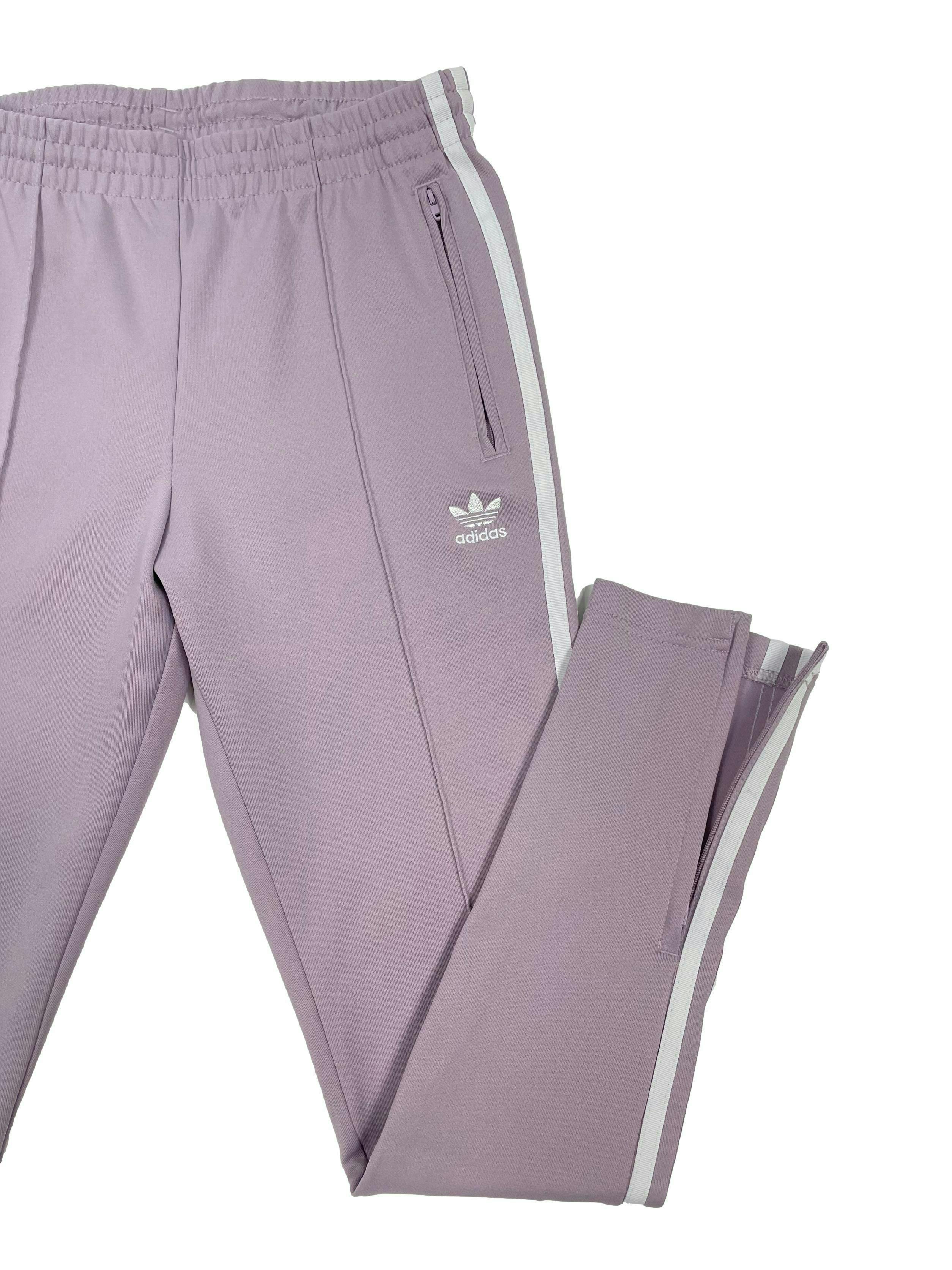 Buzo Adidas lila con líneas blancas laterales, bolsillos y bastas con cierre, pretina elástica y pasador, corte recto. Cintura 68cm Largo 91cm.
